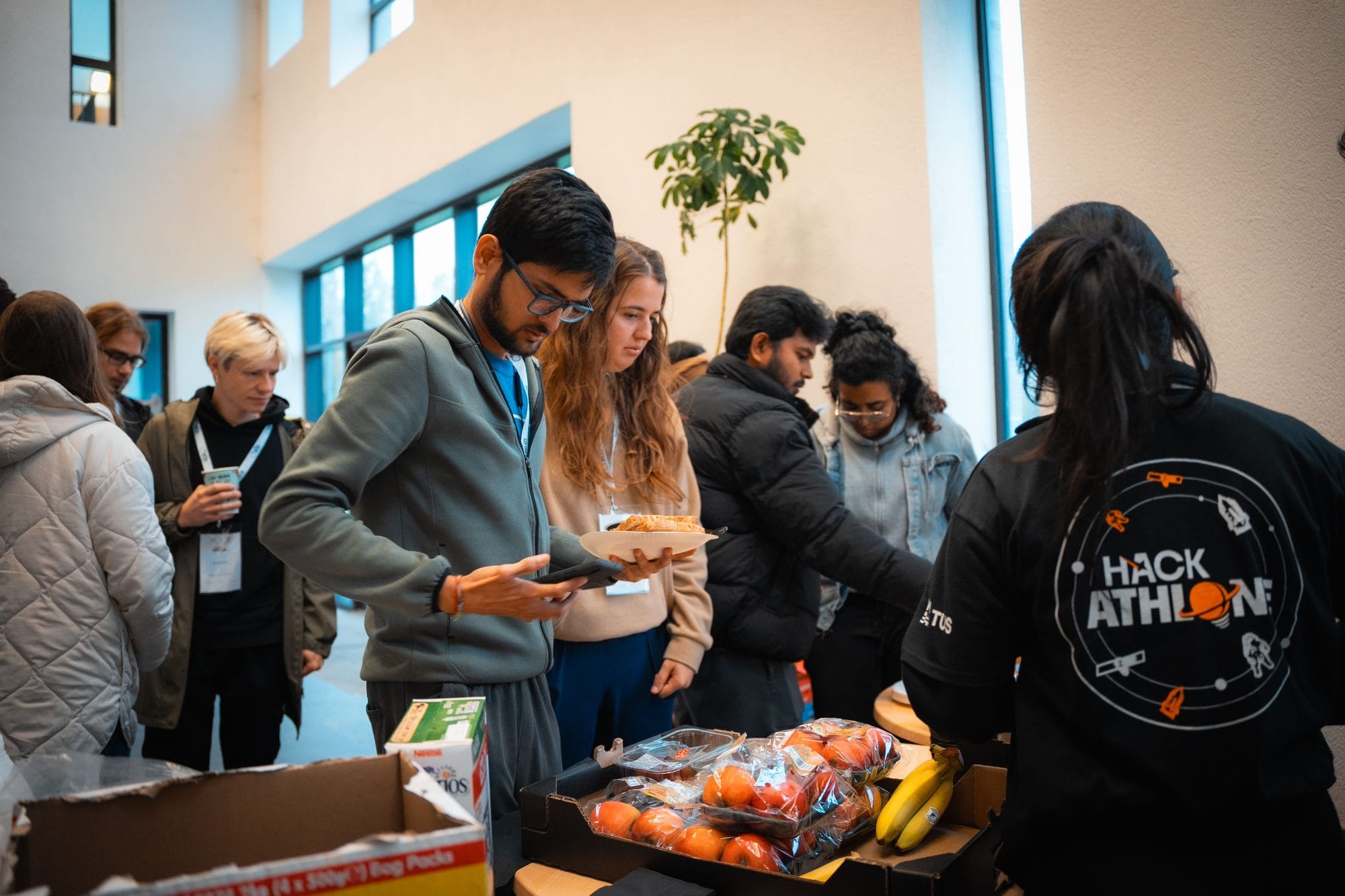 A crew member, the HackAthlone logo on their back, sets out breakfast as participants queue with plates at HackAthlone 2025