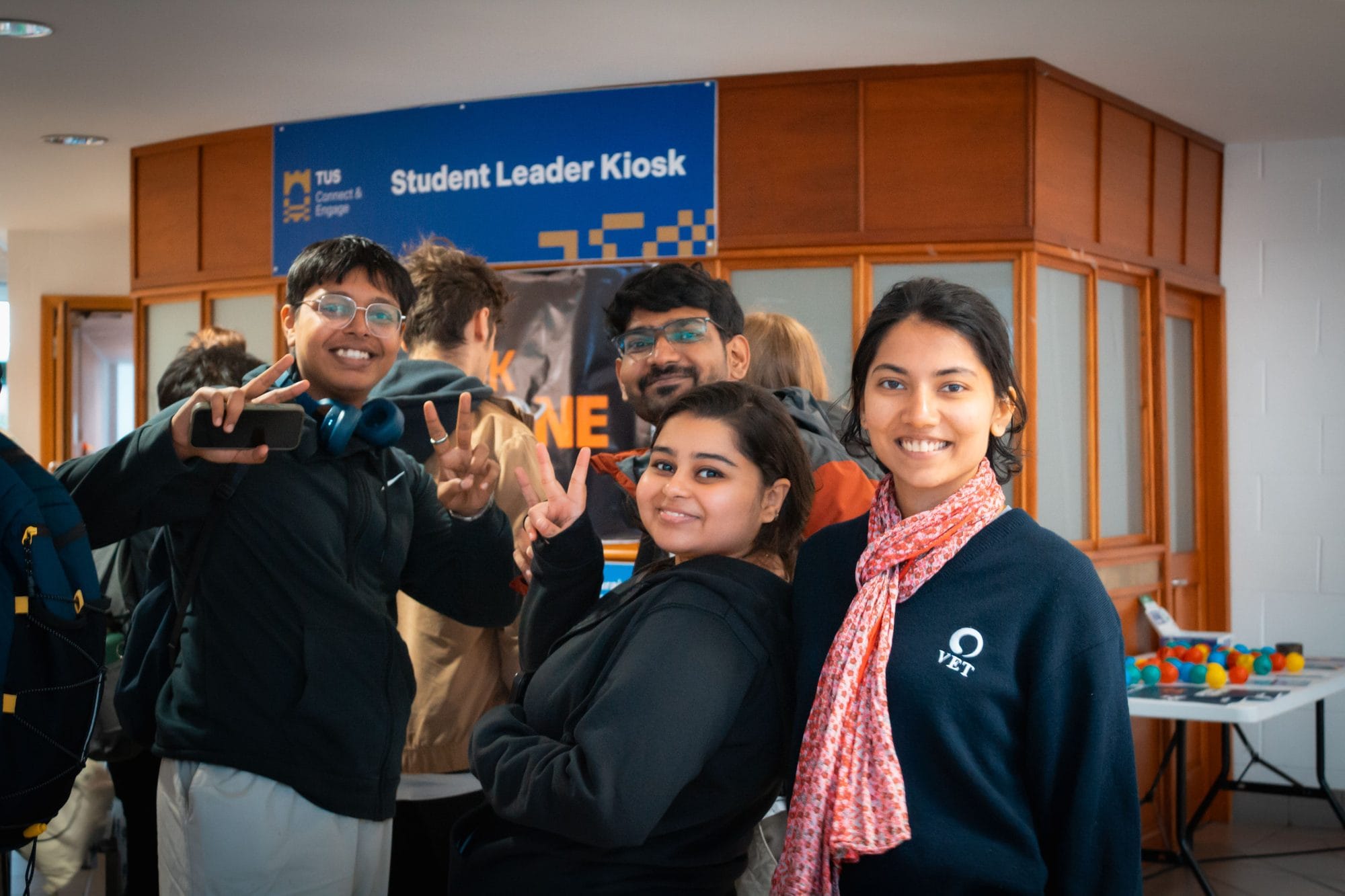 Friends smiling and making peace signs at the HackAthlone 2025 registration point, by the Student Leader Kiosk