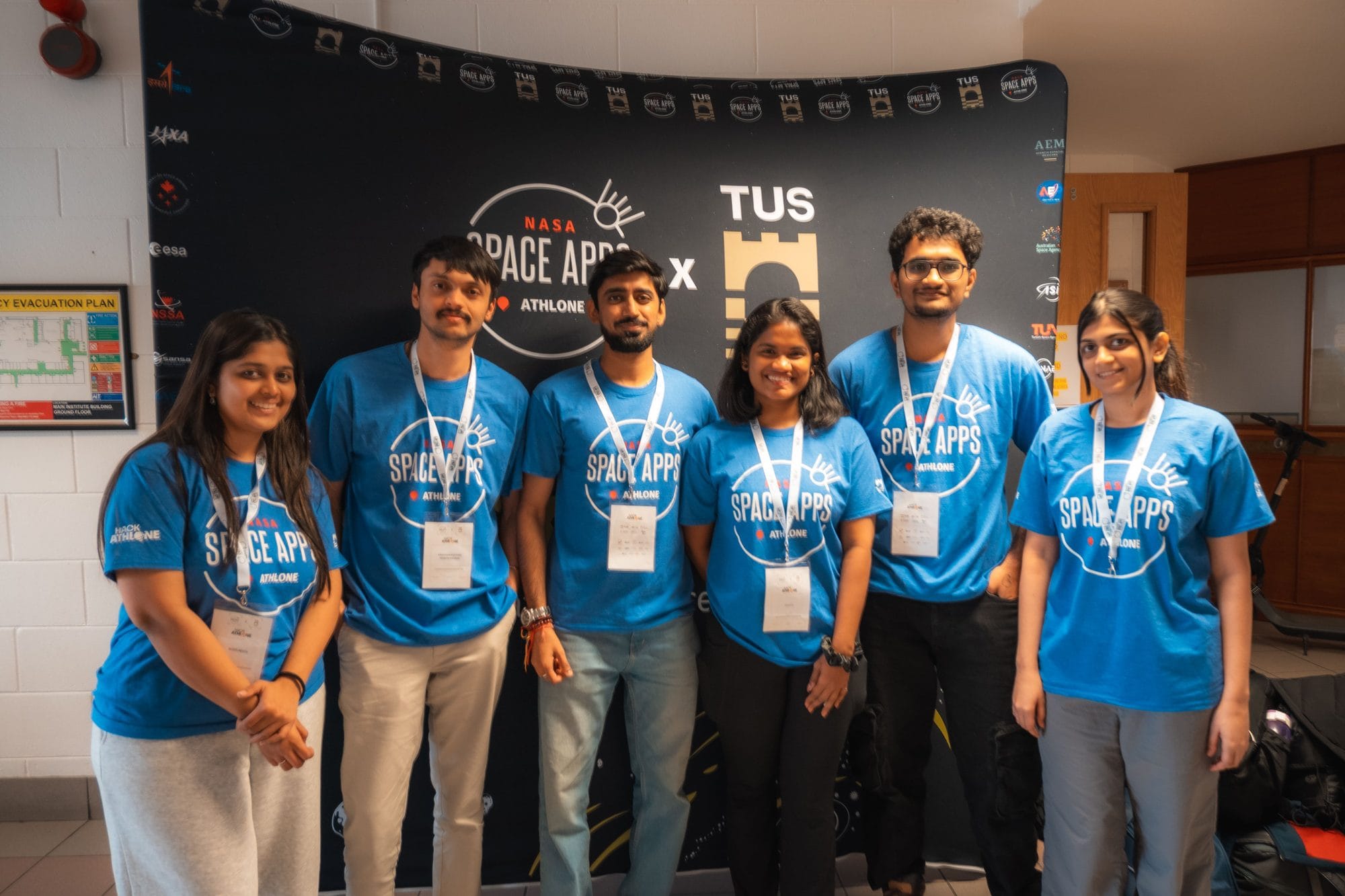 A team of six in blue NASA Space Apps Athlone T-shirts in front of the Space Apps and TUS backdrop at HackAthlone 2025
