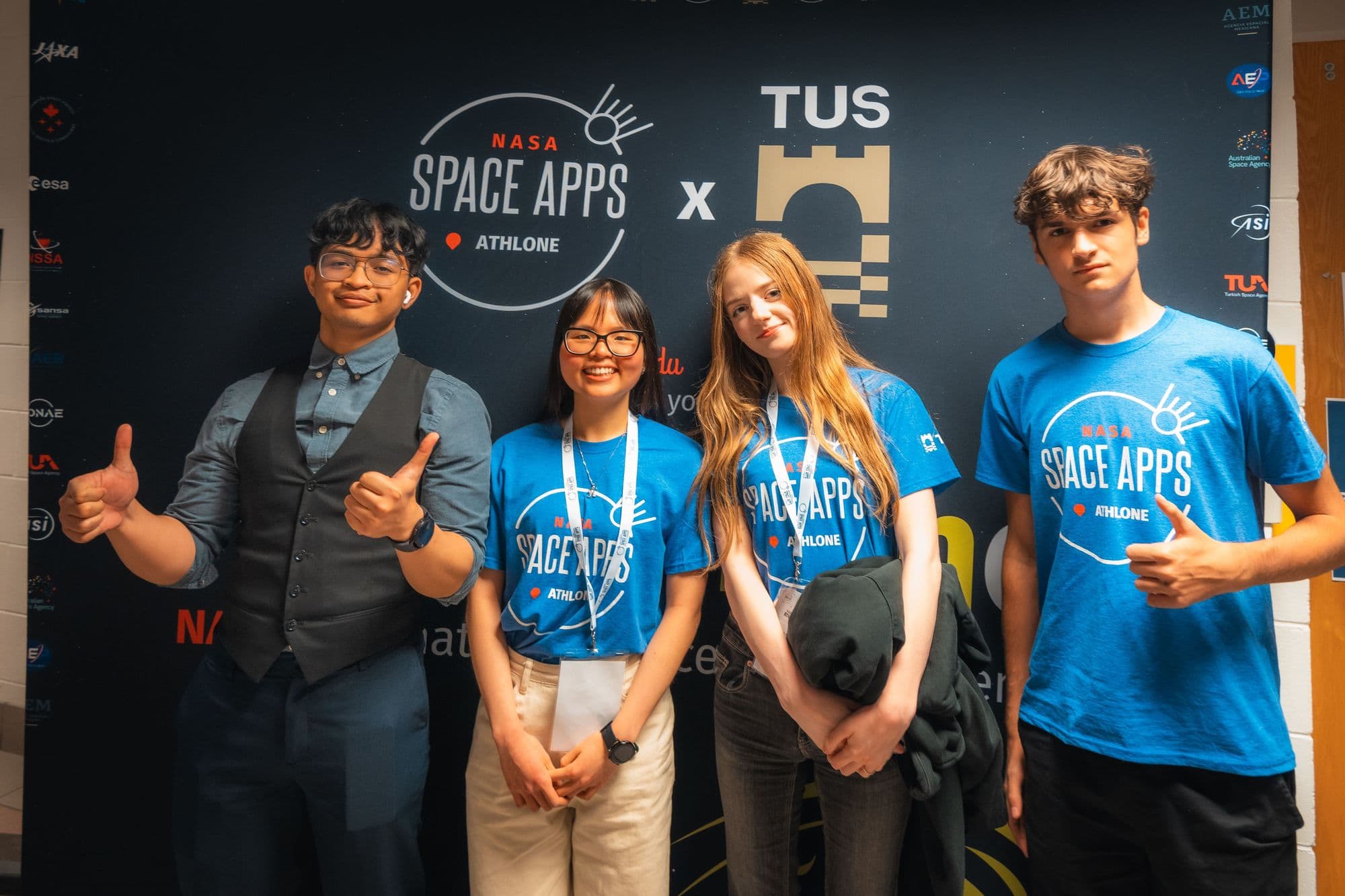 Four participants in NASA Space Apps Athlone T-shirts giving thumbs up in front of the Space Apps and TUS backdrop