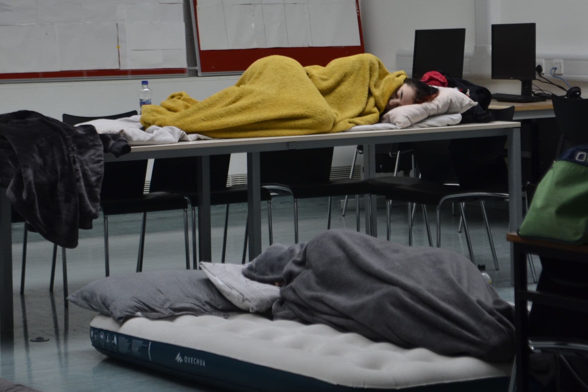 A participant asleep under a blanket on a desk, with an air mattress and sleeping bags on the floor, during the overnight at HackAthlone 2023